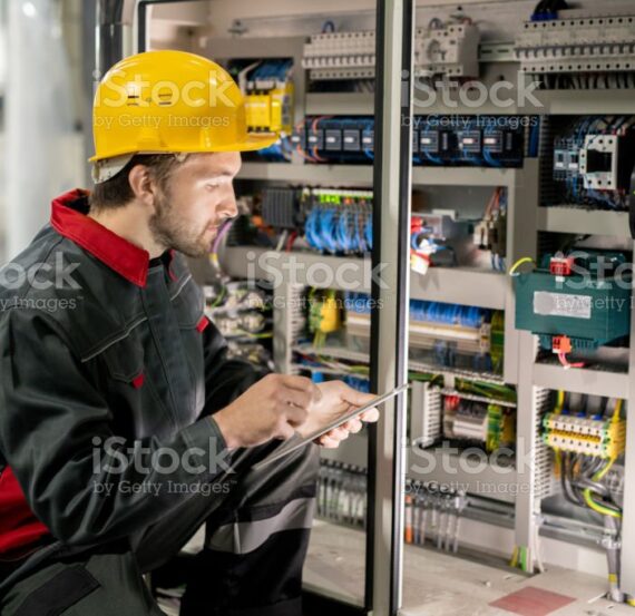 Contemporary young engineer in workwear and hardhat scrolling through data in tablet while squatting by one of industrial machines